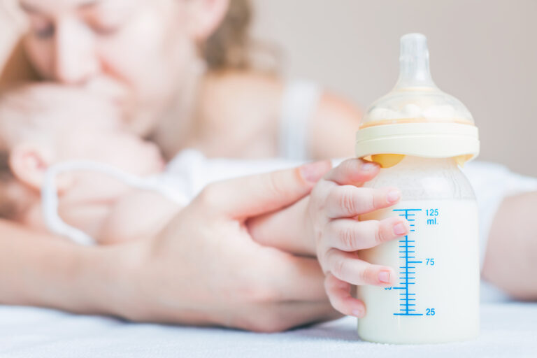 Symbolbild: Ein Baby hält ein Fläschchen und er Hand. Die Mutter ist daneben.