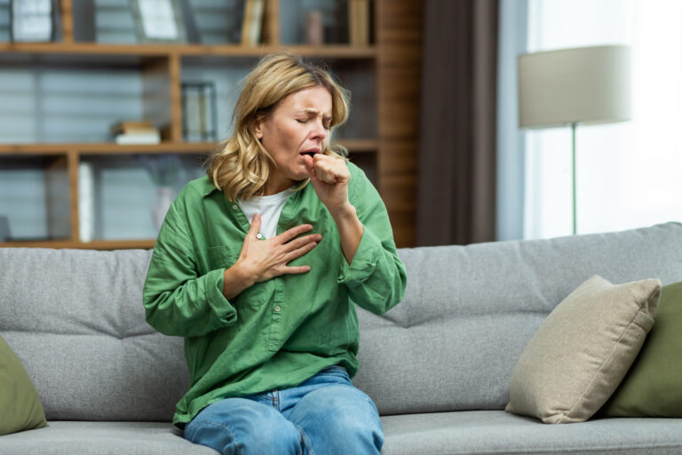 Symbolbild: Eine Frau sitzt auf einem Sofa und hustet in ihre Hand, während sie sich mit der anderen Hand an die Brust fasst.