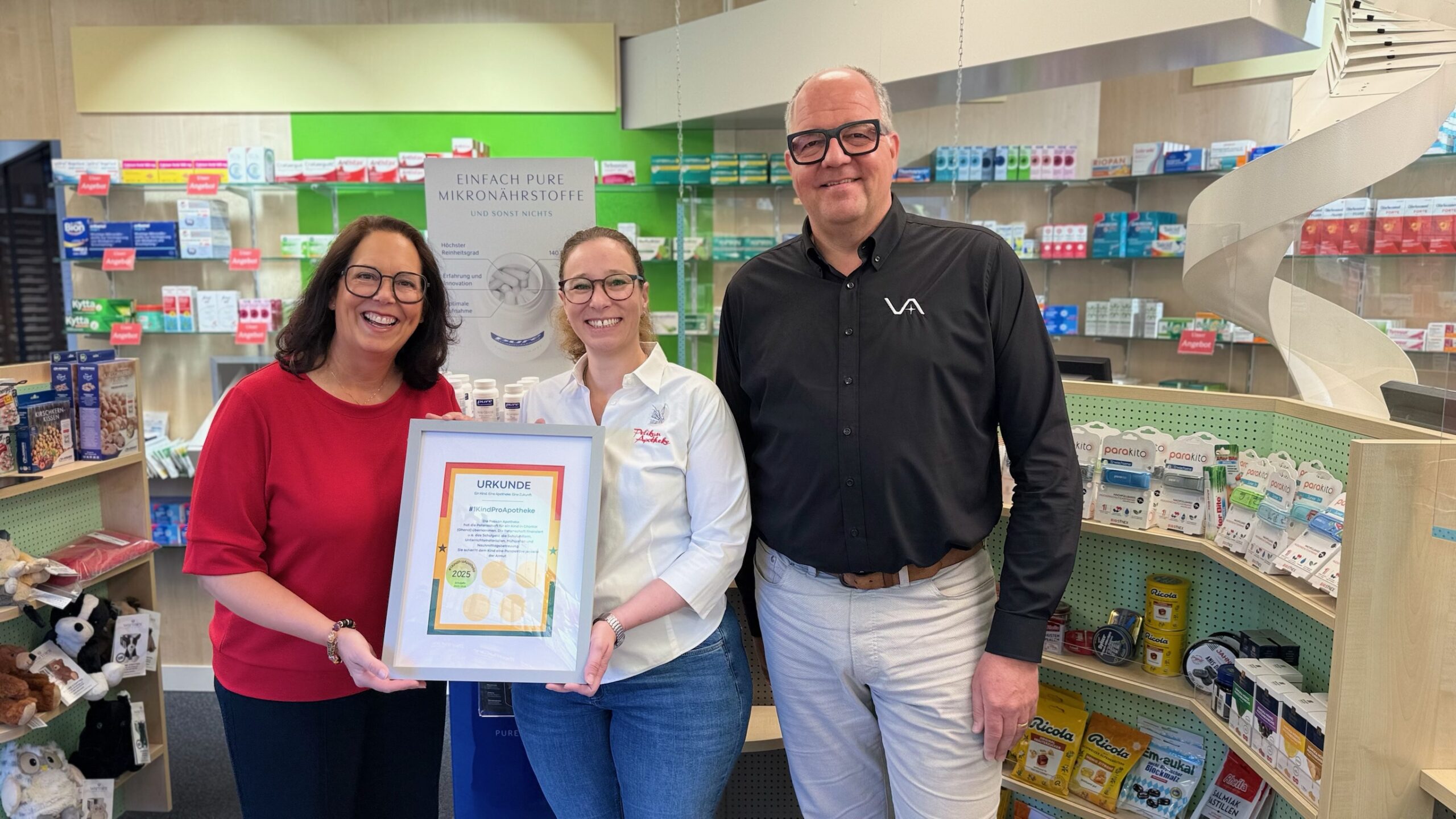 In einer Apotheke stehen zwei Frauen und ein Mann. Die Frauen halten gemeinsam eine gerahmte Urkunde in der Hand.
