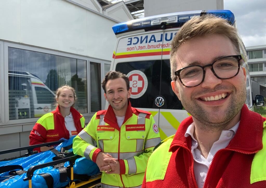Reinhard Gründler in Rettungsuniform mit seiner Schwester Viktoria und Kollegen Jakob Hager. Sie stehen von einem Rettungswagen und stützen sich auf einer Rettungstrage ab.