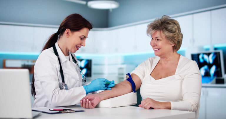 Ärztin in weißem Mantel sitzt links von einer Frau um die 70. Diese hat eine Blutspre-Binde um den Oberarm und die Ärztinn entnimmt Blut aus der Armbeuge.