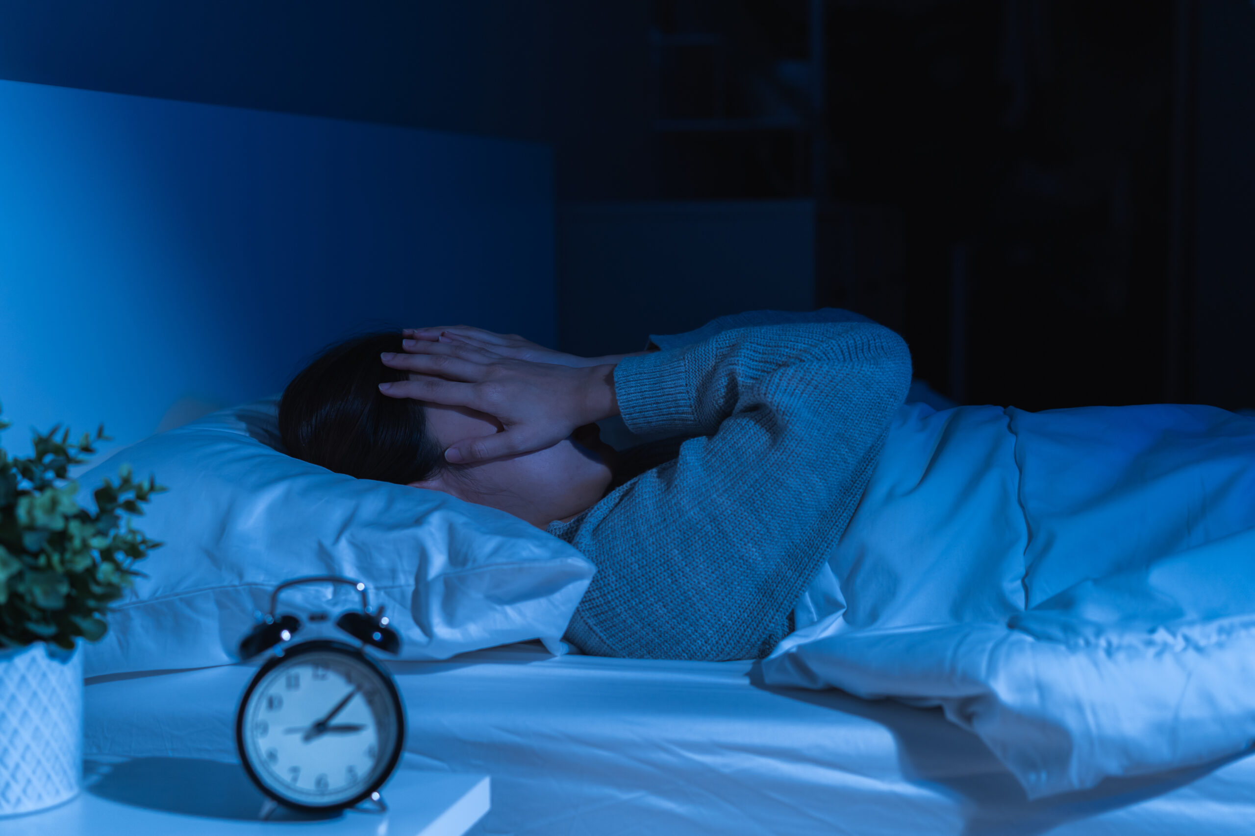 Frau liegt im Bett un dhält sich beide Hände über das Gesicht. Es ist dunkel im Raum, Die Uhr am Nachttisch zeigt 3:10.
