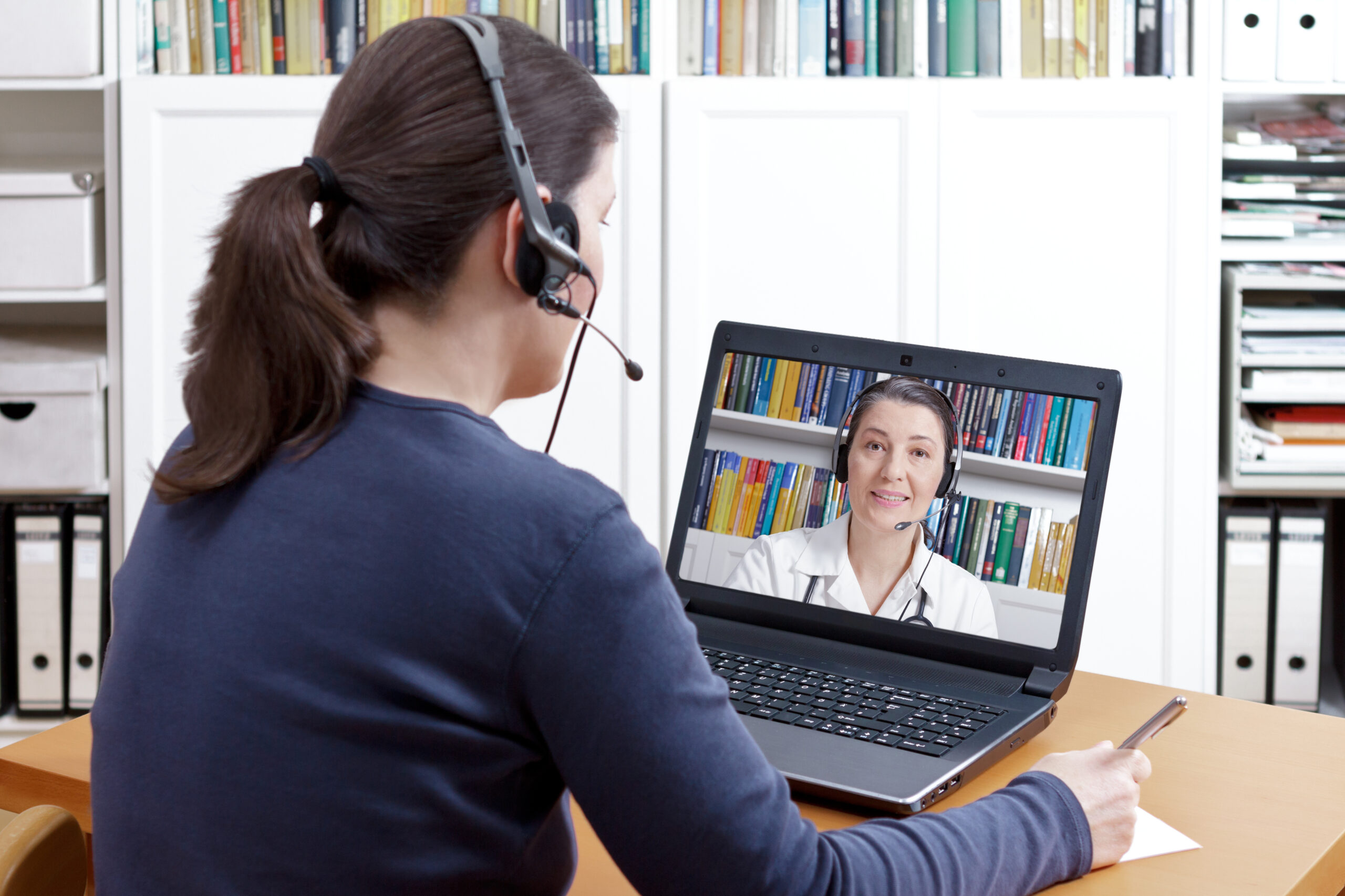 Frau sitzt mit headset vor dem Laptop. Am Bildschirm ist eine Ärztin zusehen, die ebenfalls ein Headset trägt.