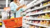 Frau steht mit Einkaufskorb vor einem Supermarktregal. In der einen Hand hält sie ein Handy und die ander hat sie nachdenklich an das Kinn gelegt.