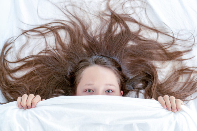 Ein Mädchen liegt im Bett, die Decke bis über die Nase hochgezogen. Seine dunklen langen Haare breiten sich fächerförmig über dem weißen Kopfpolster aus. Mit den Händen hält sie die weiße Bettdecke hochgezogen und hat die Augen offen.
