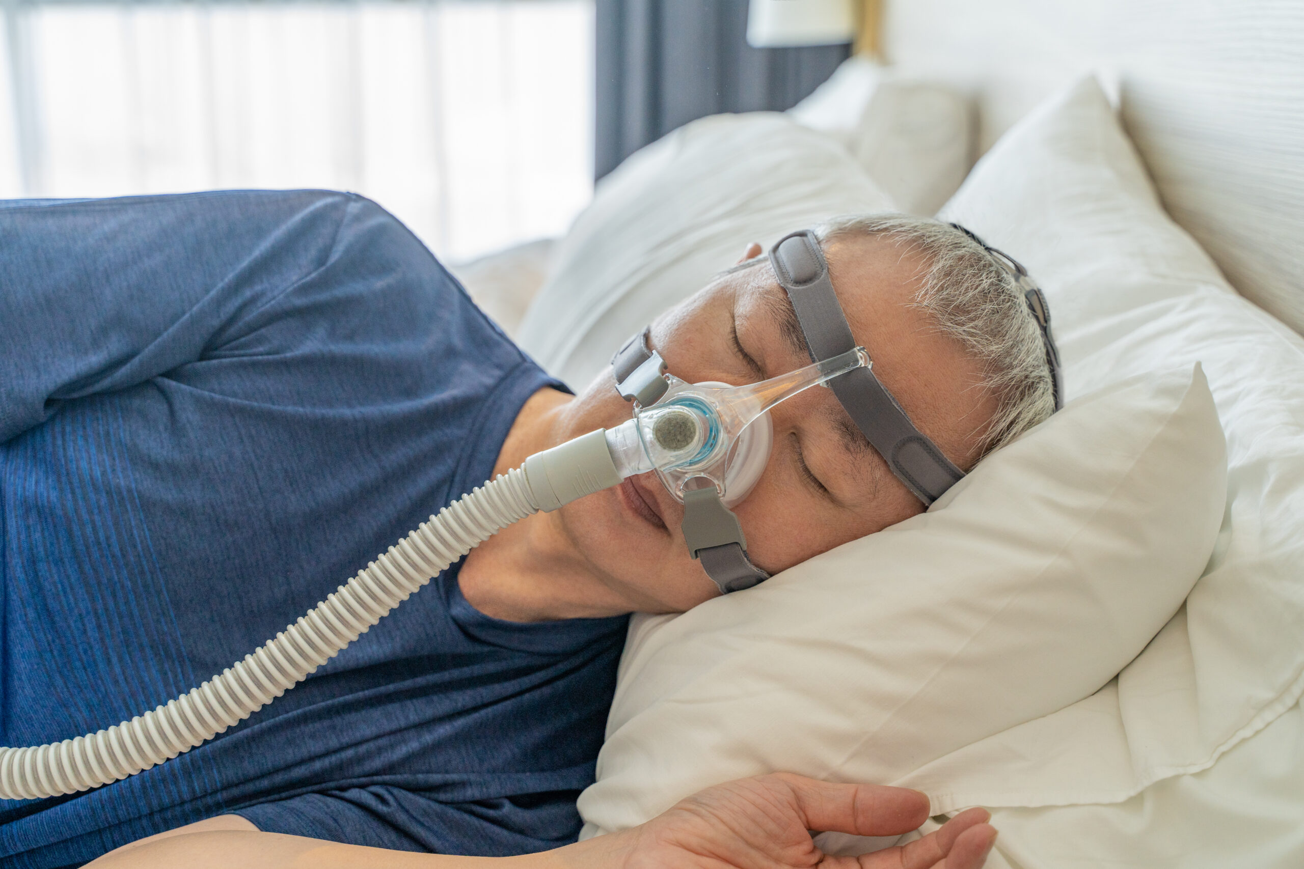Mann mittleren Alters liegt im Bett auf der Seite. Mit zwei Gurten um den Kopf ist ein Beatmungsschlauch an seiner Nase befestigt.