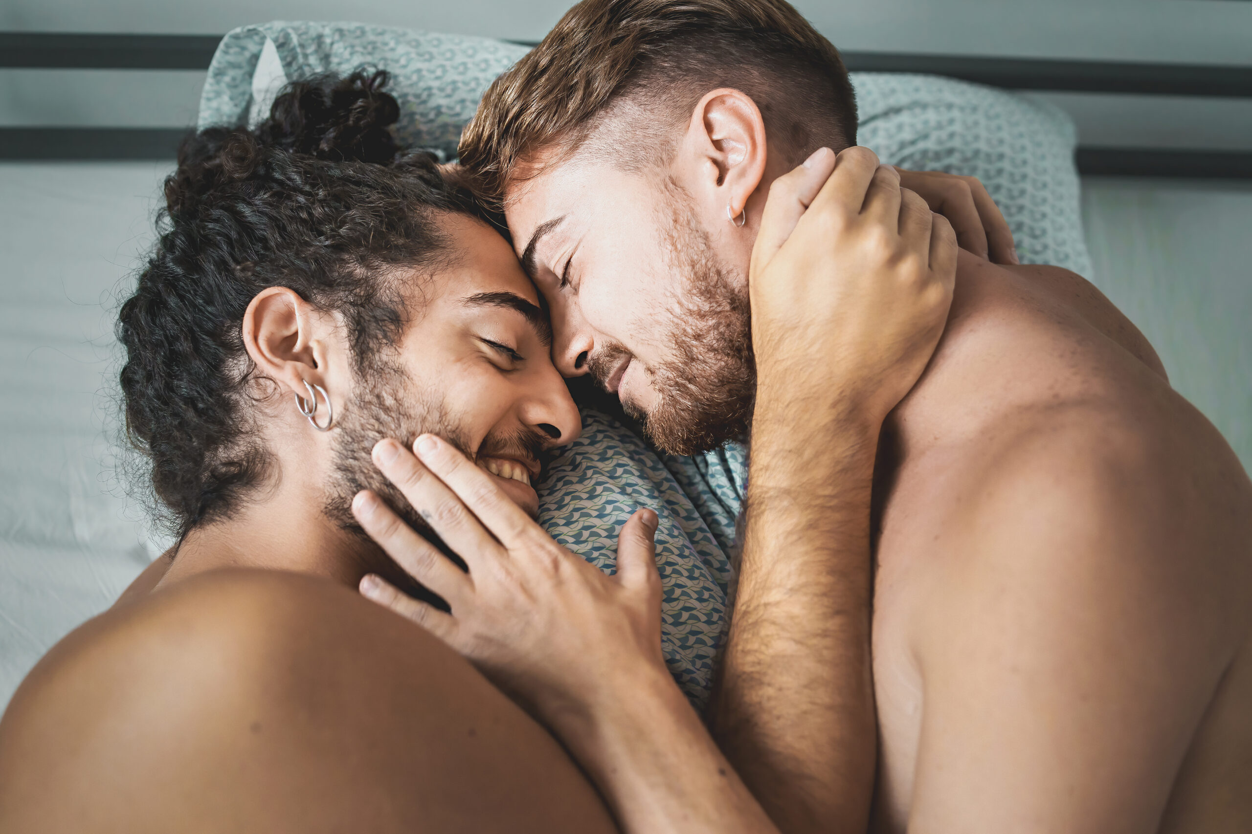 Zwei junge Männer liegen nackt im Bett. Sie umarmen sich liebevoll, lächeln leicht und haben die Stirn aneinander gelegt.