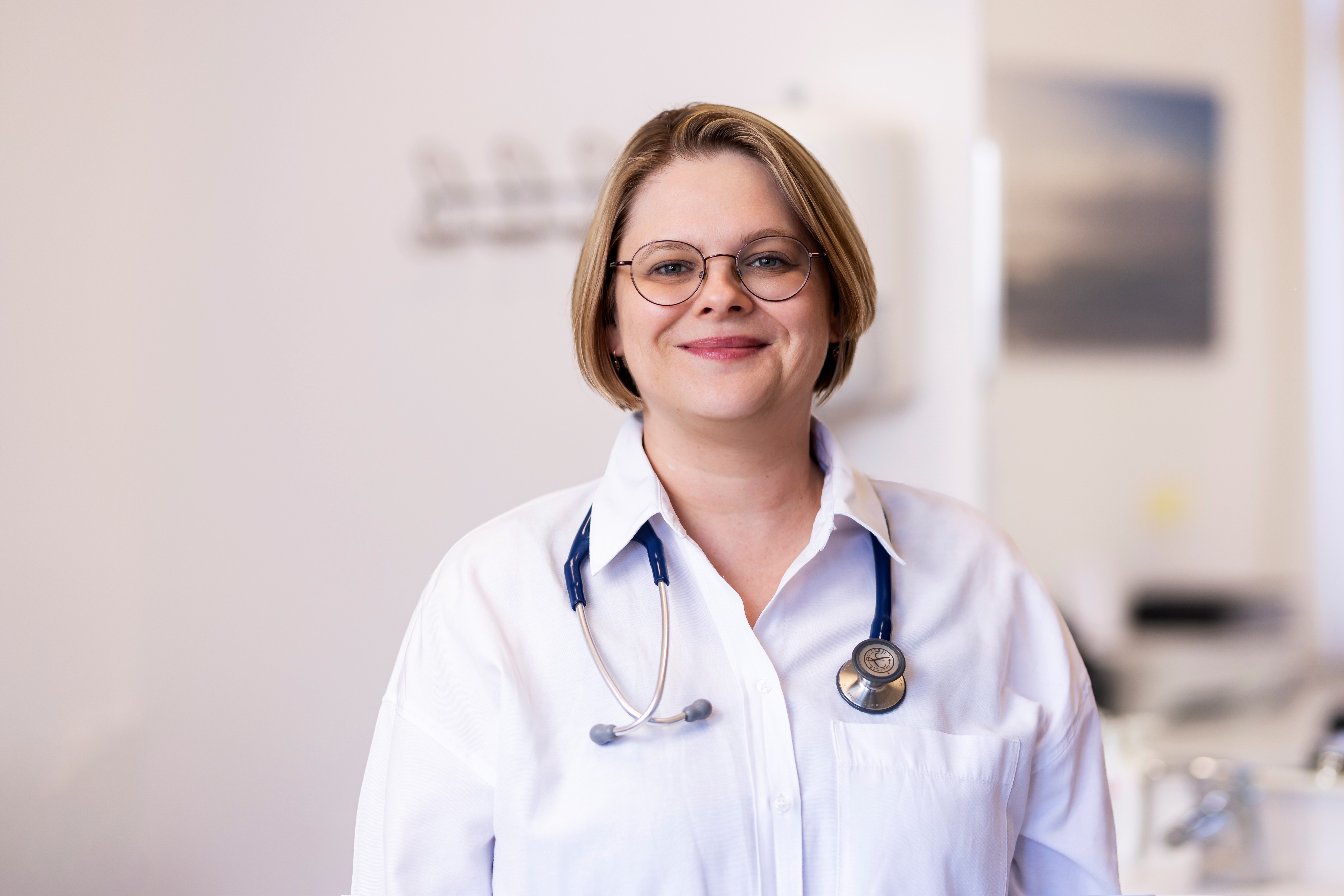 Dr. Fedra-Machacek trägt eine weiße Bluse und ein blaues Stethoskop um den Hals. Sie hat kinnlange blonde Haare, trägt eine dezente Brill und lächelt in die Kamera.