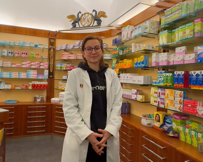 Apotheken-Influencerin Griesser steht inmitten einer Offizier von Apothekenware umringt. Sie trägt einen weißen Mantel und verschränkt die Hände.