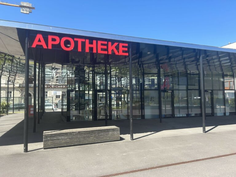 Das Glasgebäude am Grünmarkt wird von außen gezeigt. Ein rotes Apothekenschild hängt am Dach.
