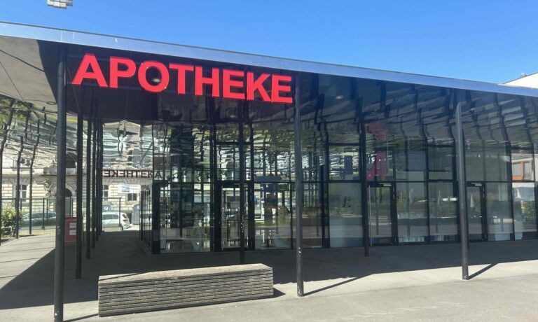 Das Glasgebäude am Grünmarkt wird von außen gezeigt. Ein rotes Apothekenschild hängt am Dach.