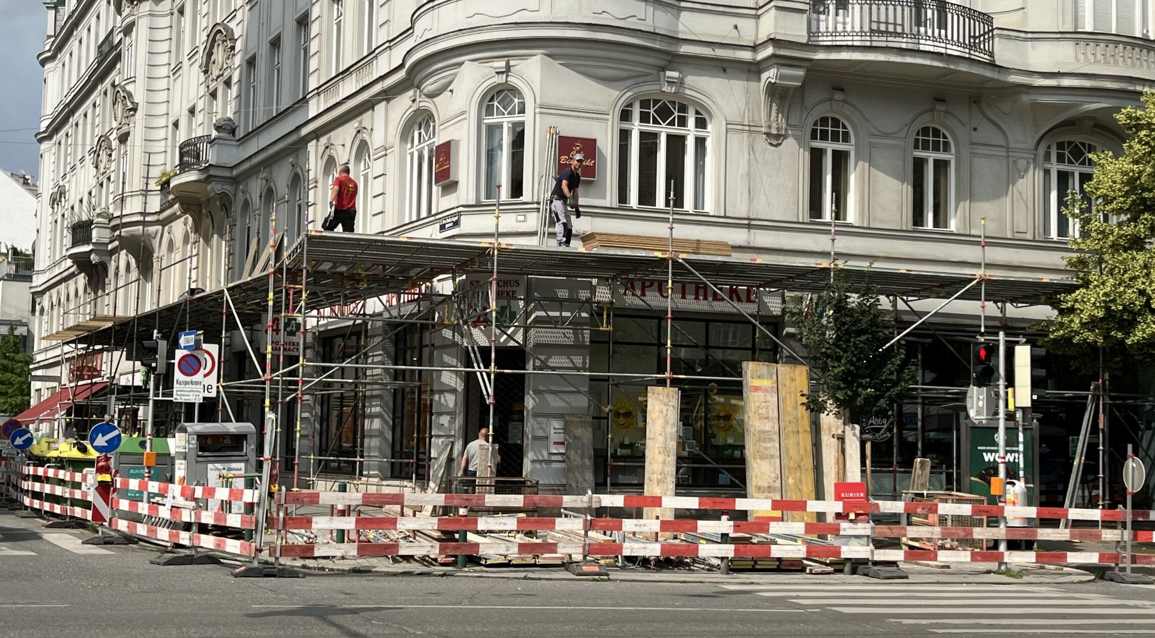 Apotheke in Ecklage mit vergitterter Eingangstüre. Davor ist der gesamte Gehsteig abgesperrt. Auf einem Gerüst sieht man Männer im Außenbeich des ersten Stocks arbeiten.