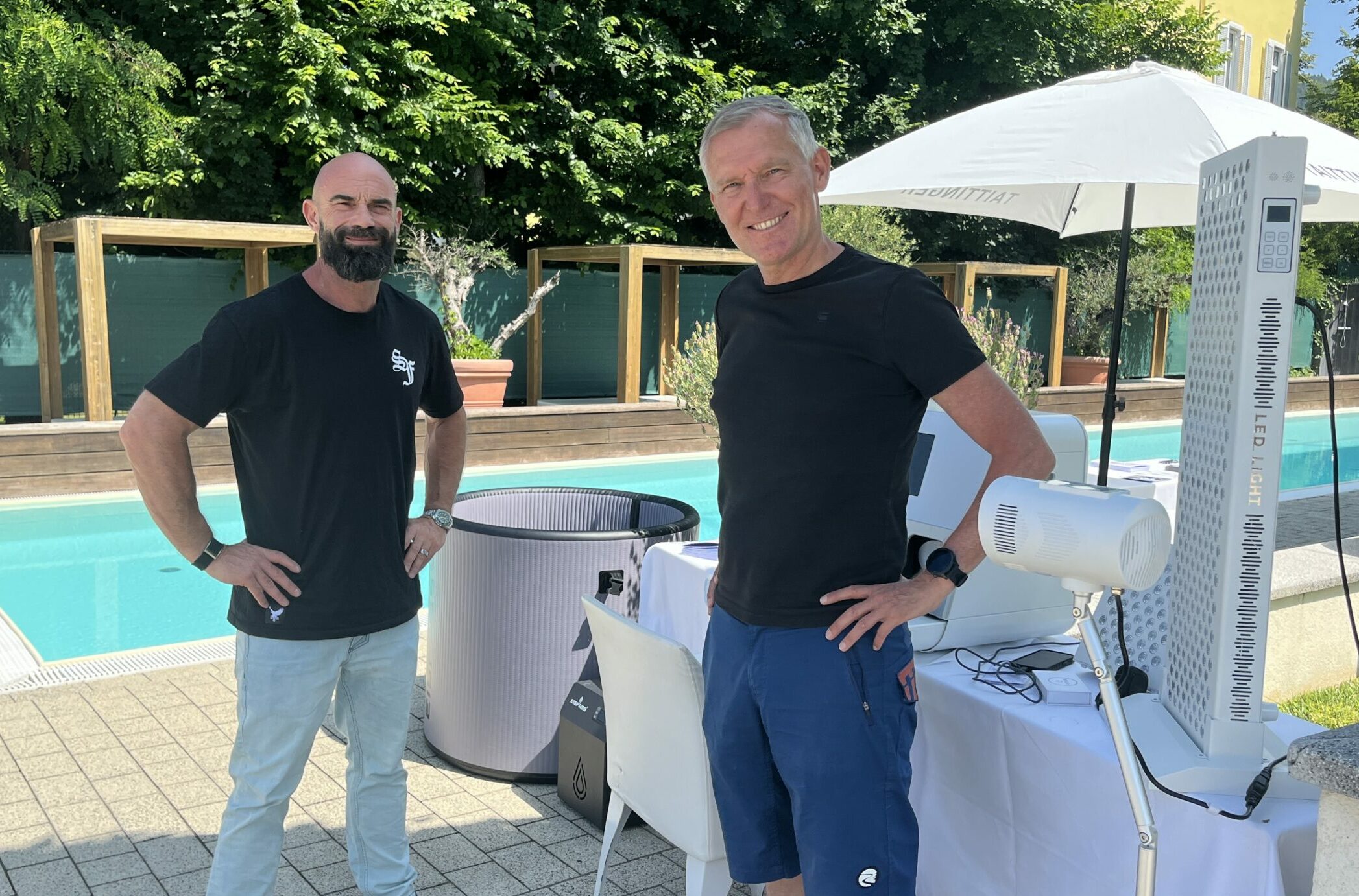 Kräftige Mann mit Vollbart und schmächtiger Mann, beide in dunkelblauen T-Shirts, stehen vor einem Pool. Direkt hinter ihnen befindet sich ein Stand mit Flyern und Messgeräten sowie eine aufblasbare, hüfthohe Tonne.