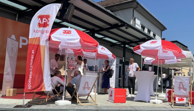 Eine Getränkebar ist verdeckte hinter rot-weißen Sonnenschirmen, weißen Liegstühlen und rot-weißen Sitzwürfeln. Vereinzelt sitzen Pesonen in den Liegestühlen und halten Gläser in der Hand.