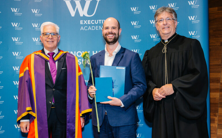 Andreas Scholz steht bei seiner Sponsion im Anzug mit Dekan und Lehrgangsleiter vor einer Fotowand der WU. Er hält sein Zertifikat in beiden Händen und eine Sonnenblume.