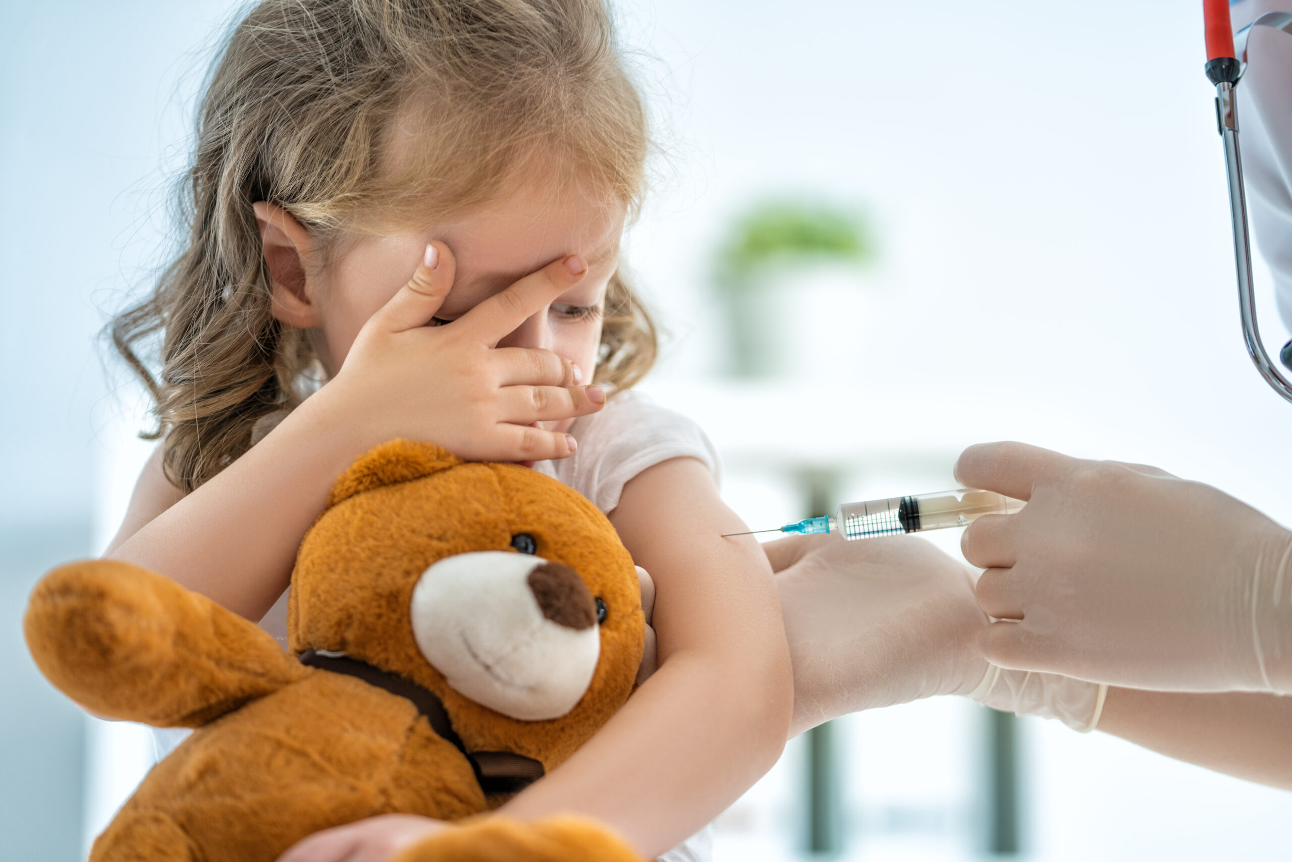 Kleines Mädchen hält einen Teddy im Arm und schaut durch die Finger auf den Arm, der gerade geimpft wird.