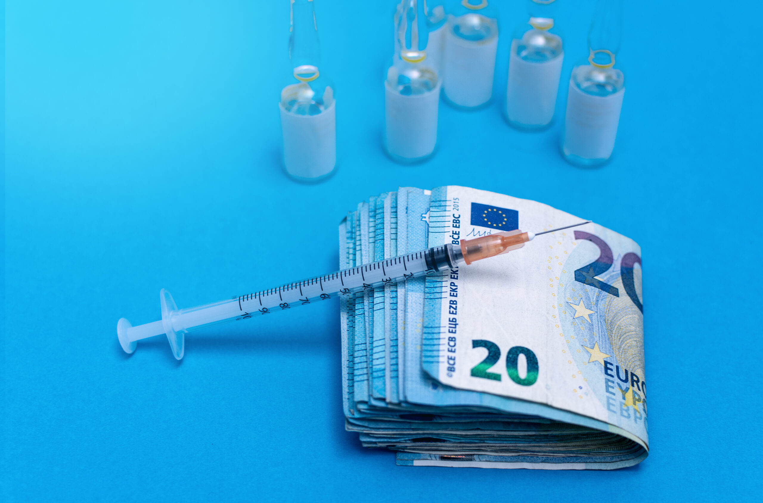 Auf dem Bild sieht man eine Spritze die auf einem Bündel 20 Euroscheinen liegt. Daneben stehen die Impfstoffflaschen.