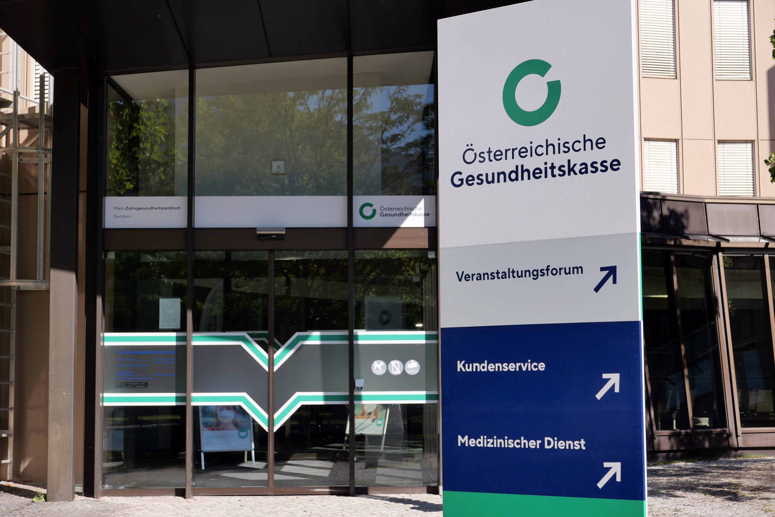 Eingang einer ÖGK-Stelle. Vor der Türe steht eine Säule mit Logo und Orientierungswegweiser.