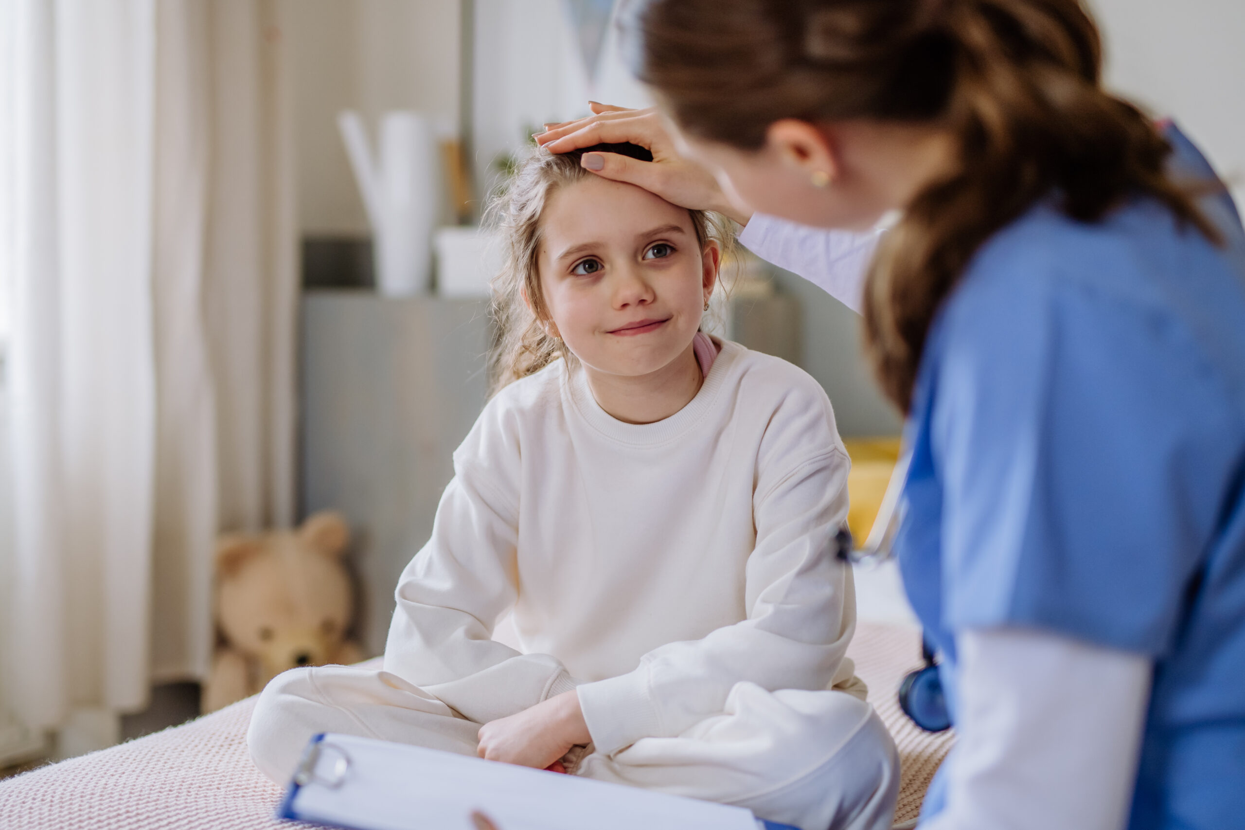 Mädchen sitzt im Pyjama im Krankenhausbett. Ärztin streichelt ihr mit dem Daumen über die Stirn.