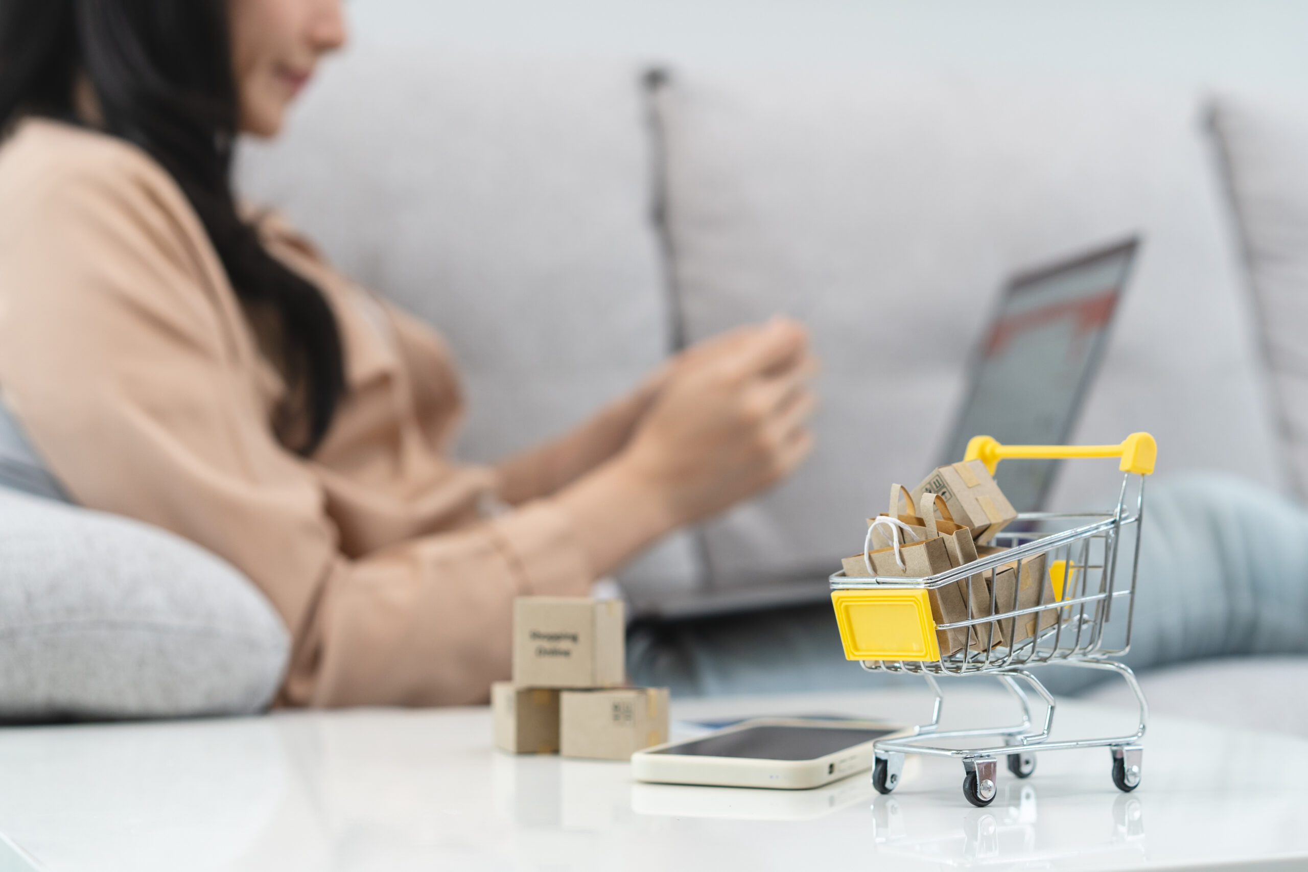 Frau sitzt auf der Couch vor ihrem Laptop. Am Tisch daneben steht ein Miniatur-Einkaufswagen mit Miniatur-Einkaufstüten.