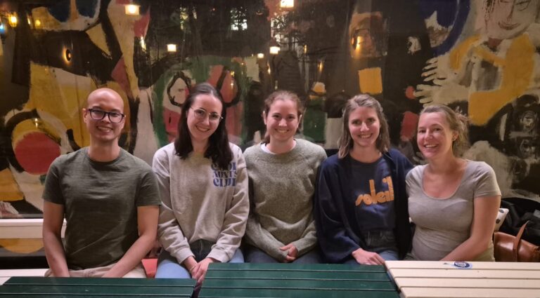 Auf dem Bild sitzen fünf junge Pharmazeut:innen an einem Tisch. Sie schauen in die Kamera und lächeln.