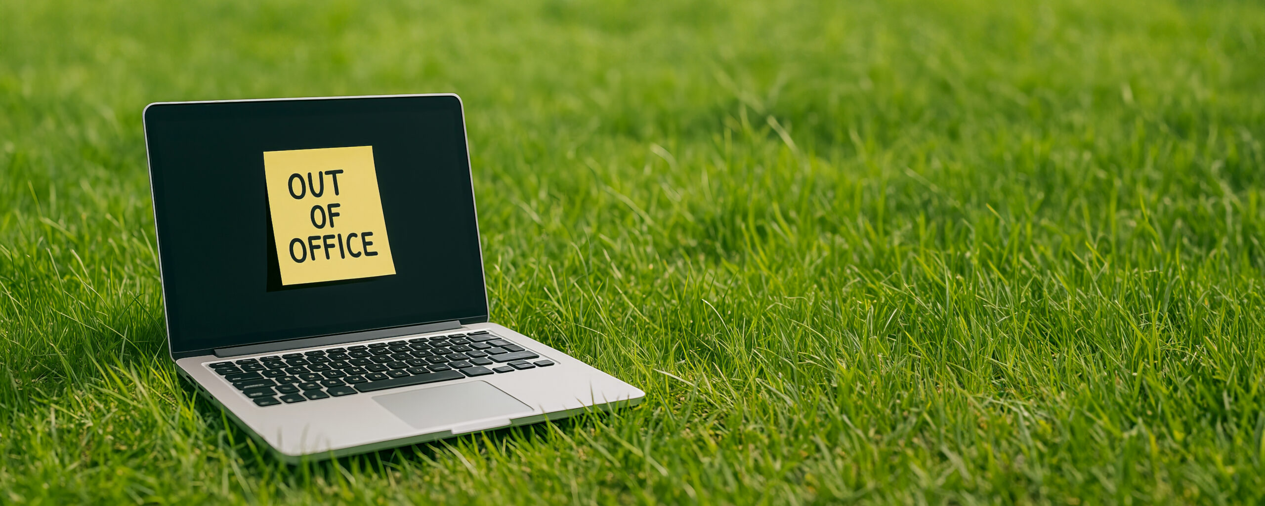 Auf einem grünen Rasen steht ein Laptop. Auf seinem geöffneten Bildschirm klebt ein Post it mit den Worten "out of office".