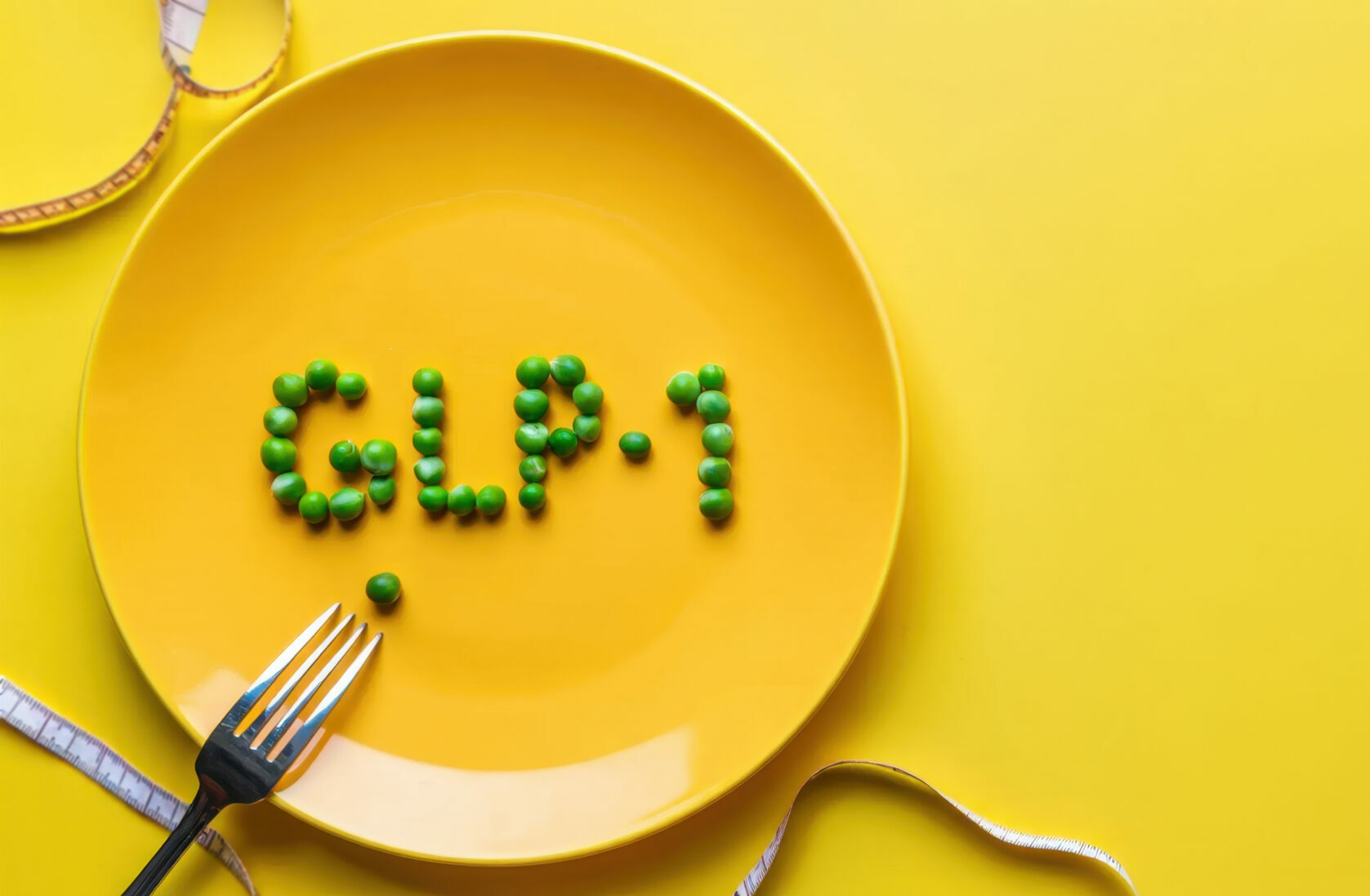 Symbolbild: auf einem gelber Teller liegen grüne Erben die das Wort "GLP-1" formen. Daneben liegt ein Maßband.