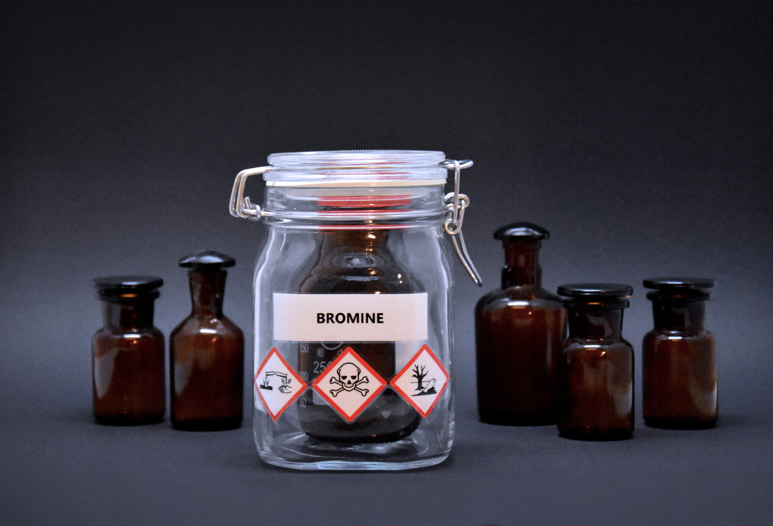 Eine Flasche Brom steht in einem Sicherheitsgefäß und ist mit Aufklebern versehen. Sie zeigen die Toxizität, ätzend und Naturschädlichkeit von Brom. Ringsum stehen braune Flaschen.