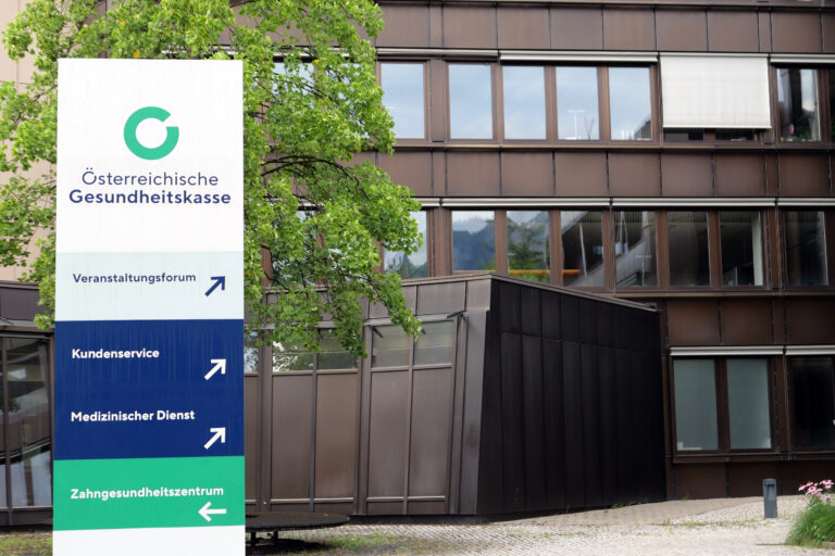 Vor einem dunkelbraunen Häuserteil steht ein weißes Schild. Auf ihm steht "Österreichische Gesundheitskasse". Es verweist auf die verschiedenen Dienste. Zum Beispiel das "Kundenservice".