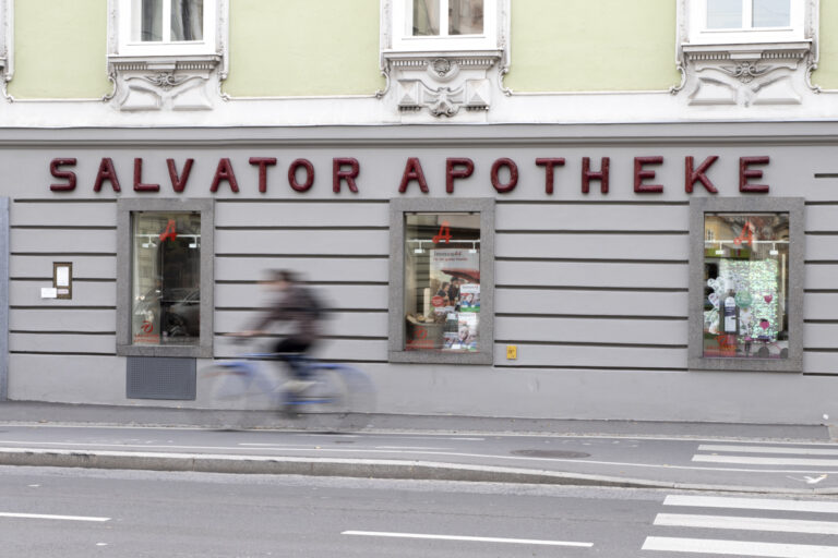 Die Salvator Apotheke liegt an einem Fahrradweg. Auf diesem fährt ein Fahrradfahrer schnell. Man sieht ihn nur verschwommen. Hinter ihm ist das graue Gebäude mit der roten Aufschrift "Salvator Apotheke".