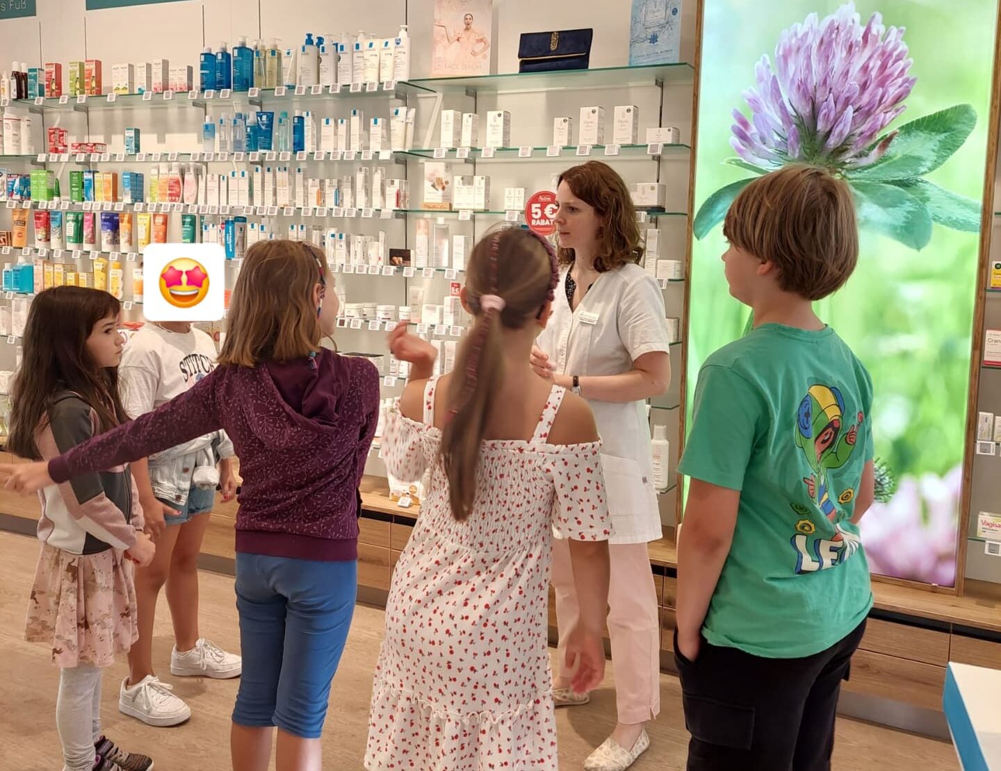Eine Gruppe Kinder steht mit einer Apothekerin in Kreis. Sie stehen in der Offizin vor einer Kosmetika-Wand.