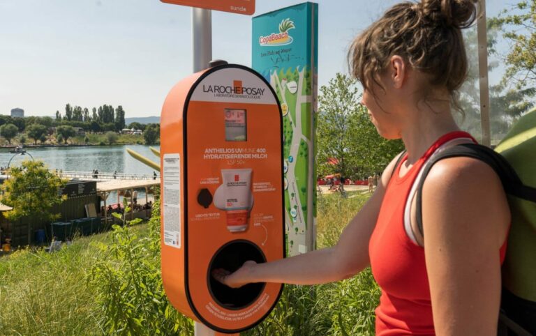 Der orangene obale Sonnencreme-Spender steht auf der Donauinsel. Eine junge Dame hält, ebenfalls in orange gekleidet, ihre Hand in das dafür vorgesehene Loch. Dort kommt eine vornotierte Menge Sonnencreme heraus.