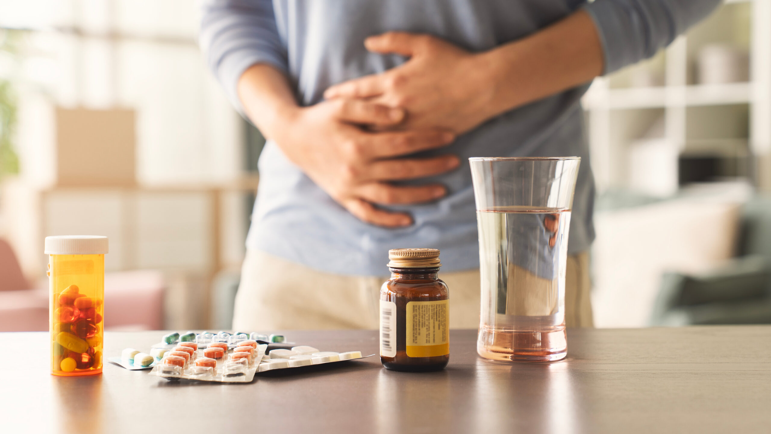 Verschiedene Tabletten und Wasserglas vor einem Mann, der sich den Bauch hält