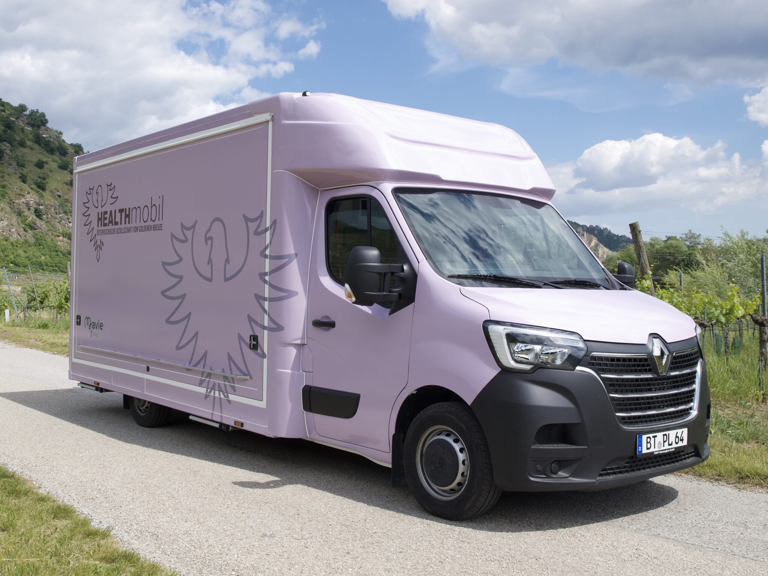 Das violette Health Mobil von der ÖGGK fährt auf einer Straße in der Natur. Man sieht wie groß es ist und dass es gut mobil unterwegs ist.