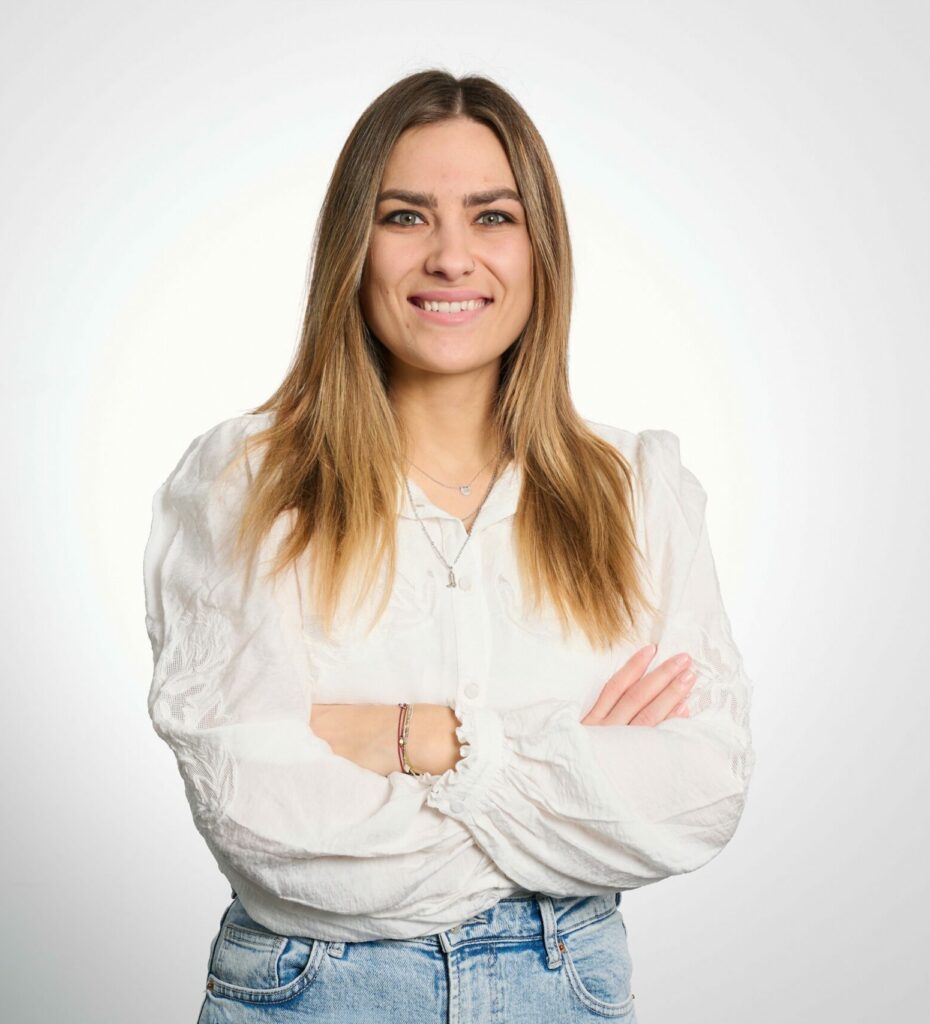 Saskia Luksch verschränkt die Arme und lacht. Sie trägt eine weiße Bluse und hat lange blonde Haare.