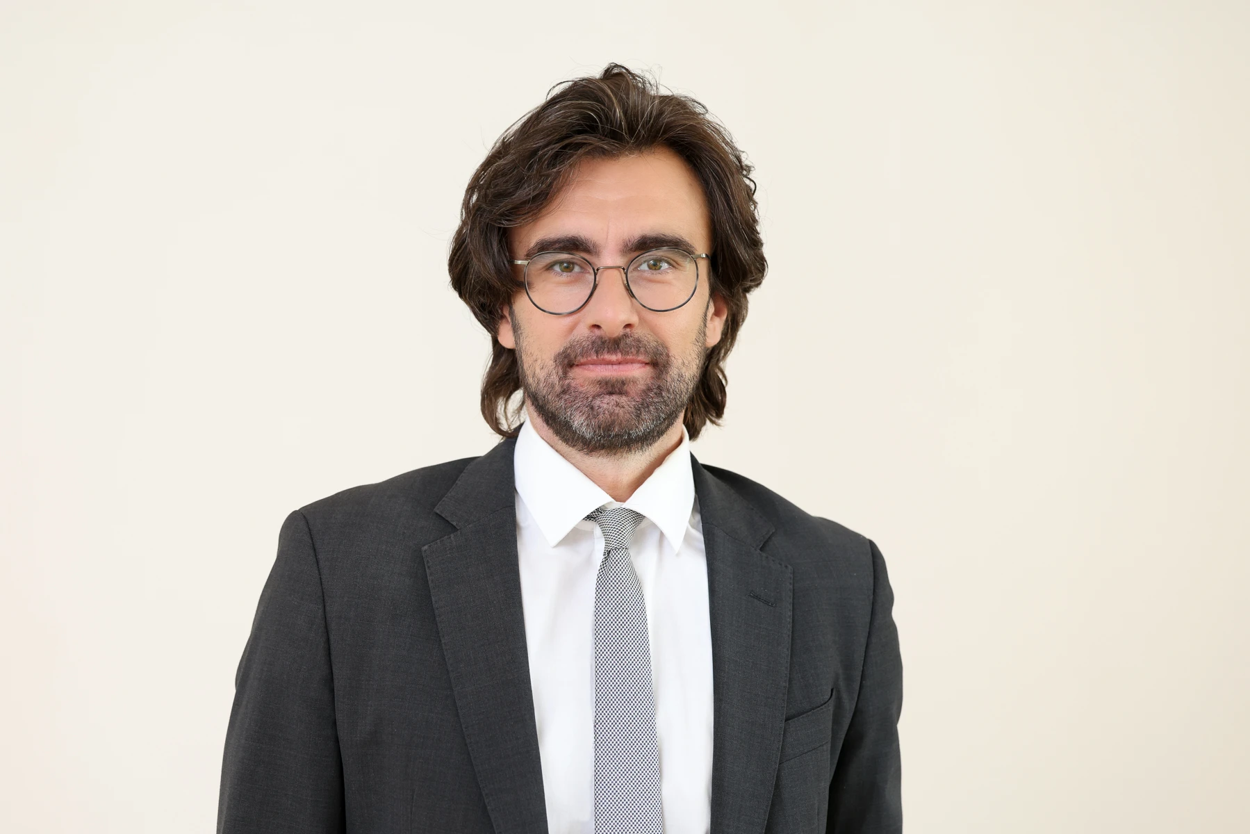 Dr. Thomas übernimmt die neue Stelle. Er hat dunkles Haar. Trägt eine Brille und einen schwarzen Anzug mit einer hellgrau-blauen Krawatte.