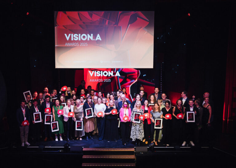 Alle Gewinner der VISION.A Awards wurden auf der Bühne zum Abschlussfoto versammelt. Sie strahlen und lachen und halten ihre Awards.