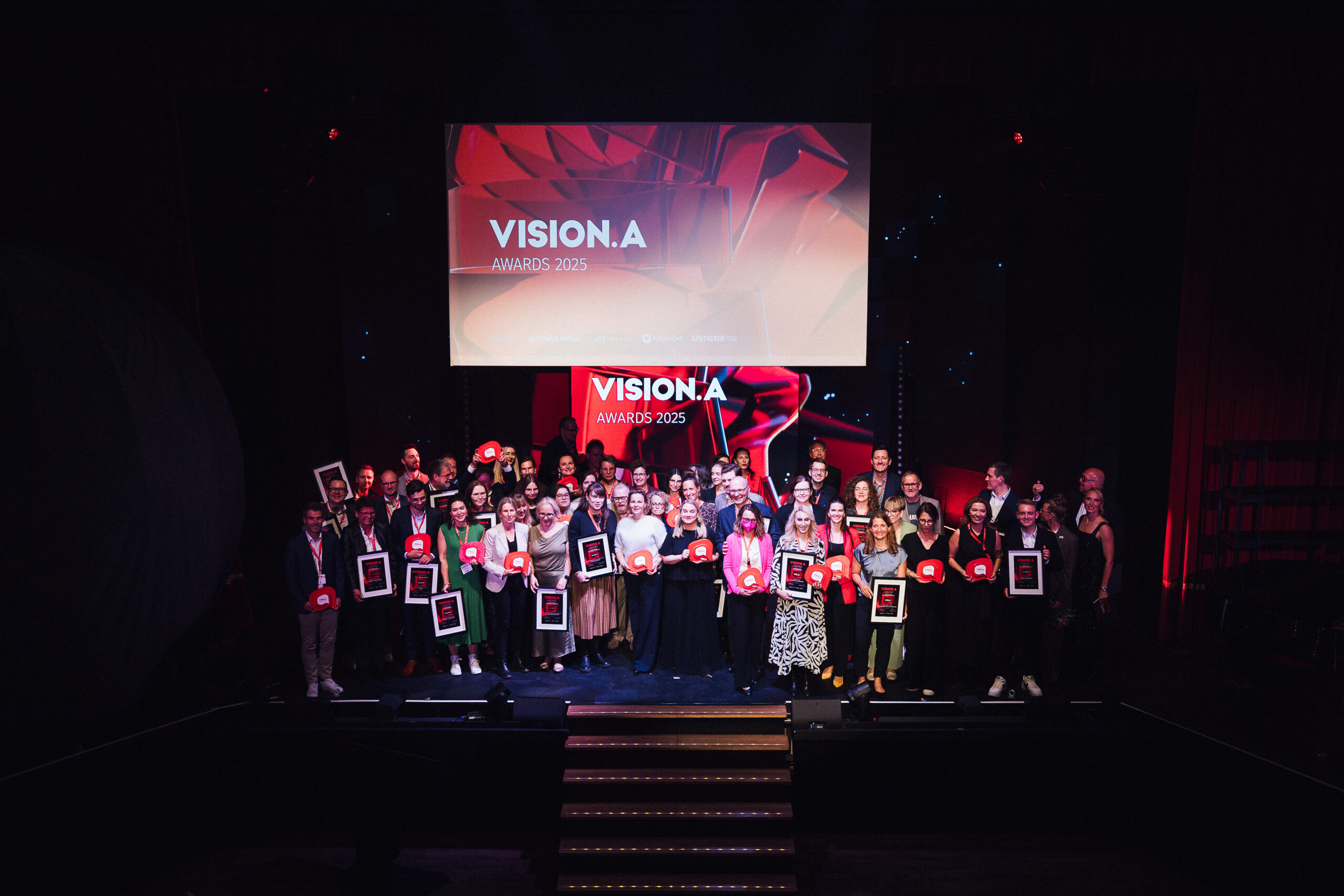 Alle Gewinner der VISION.A Awards wurden auf der Bühne zum Abschlussfoto versammelt. Sie strahlen und lachen und halten ihre Awards.