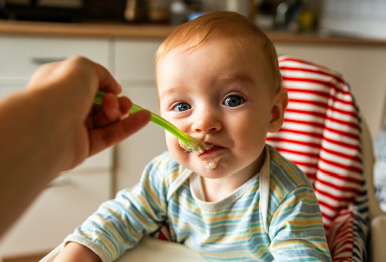 Symbolbild: Ein kleines Kind sitzt im Hochstuhl und bekommt Beikost gefüttert. Es ist um den Mund mit Brei verschmiert.