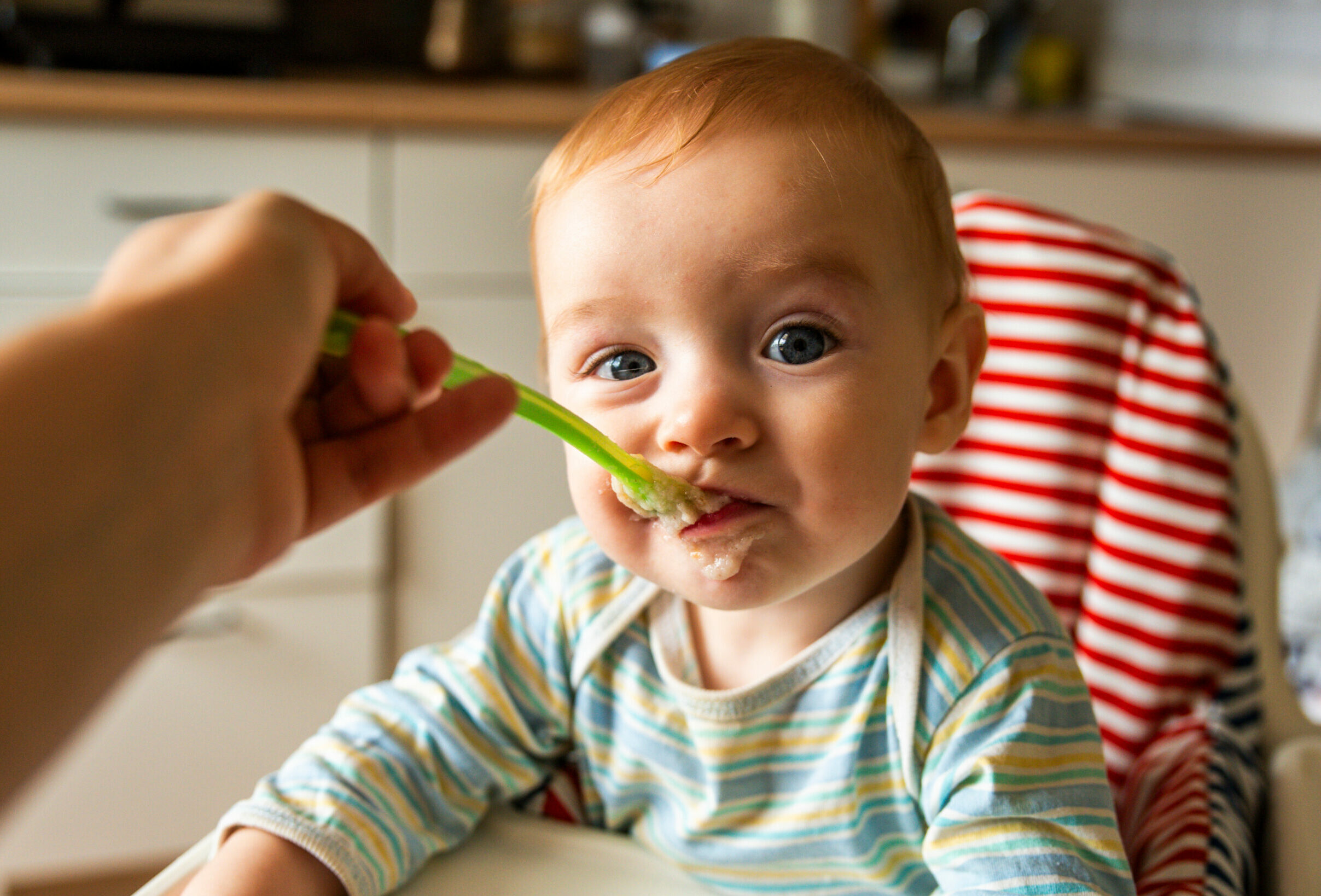 Symbolbild: Ein kleines Kind sitzt im Hochstuhl und bekommt Beikost gefüttert. Es ist um den Mund mit Brei verschmiert.