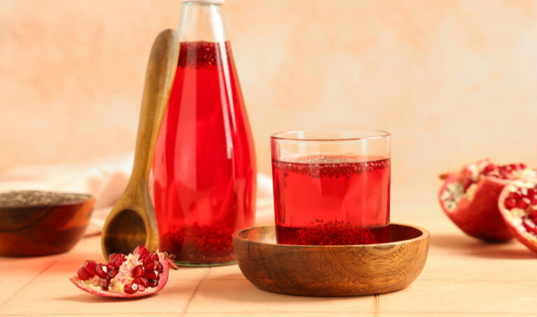 Symbolbild: Eine Glasflasche mit roten Granatapfelsaft steht auf einem Tisch. Vor ihr liegen Teile eines Granatapfels und eine Holzschale.