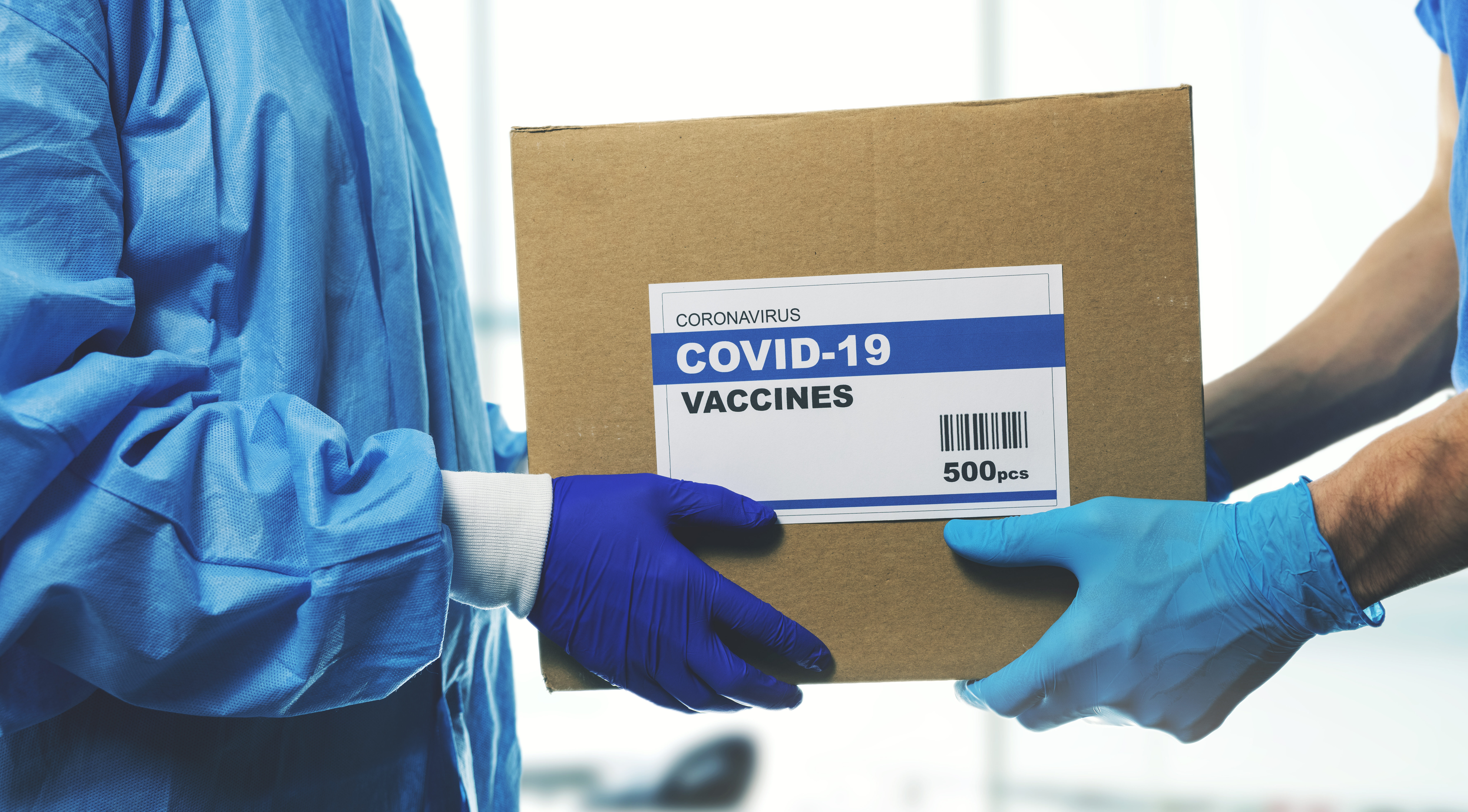 Symbolbild: Ein Karton mit der Aufschrift "Corona Impfungen" wird von einer Person zur anderen übergeben. Die linke Person trägt einen blauen Overall und Gummihandschuhe. Die Rechte hat blaue Gummihandschuhe.