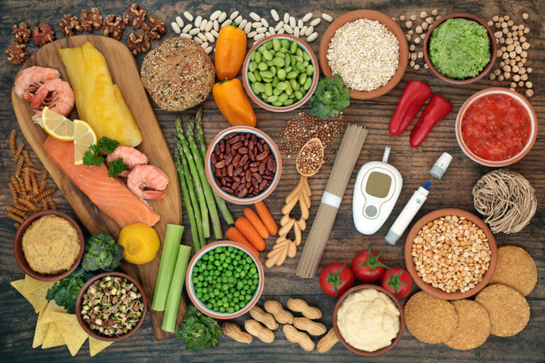 Symbolbild: Viele verschiedene Früchte, Obst und Nüsse liegen auf einer Tischplatte. Mitten drinnen liegt ein Blutzuckermessgerät und eine Stechhilfe.