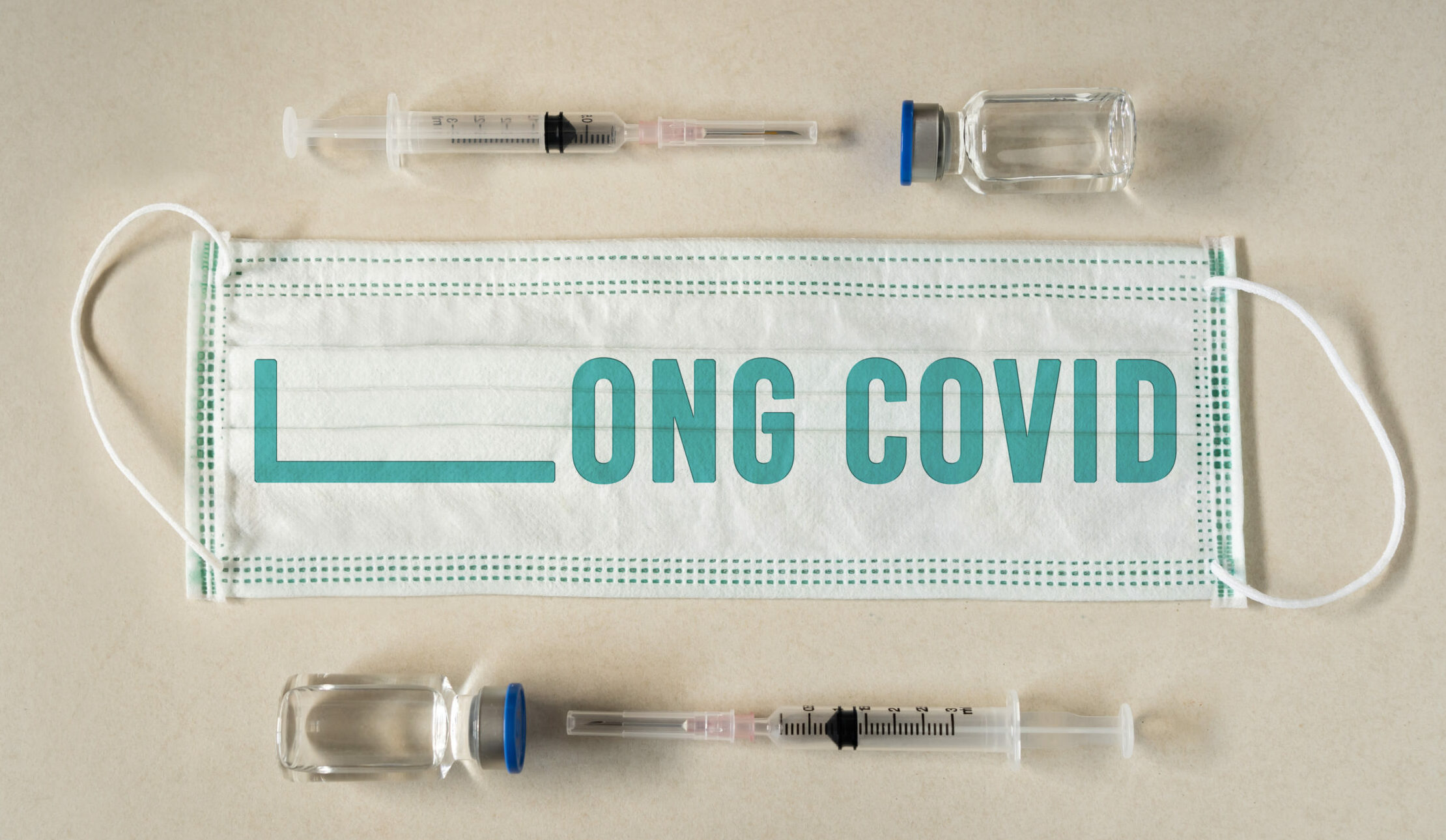 Symbolbild: Ein Mund Nasenschutz liegt neben Impfungen und auf ihm steht "Long COVID".