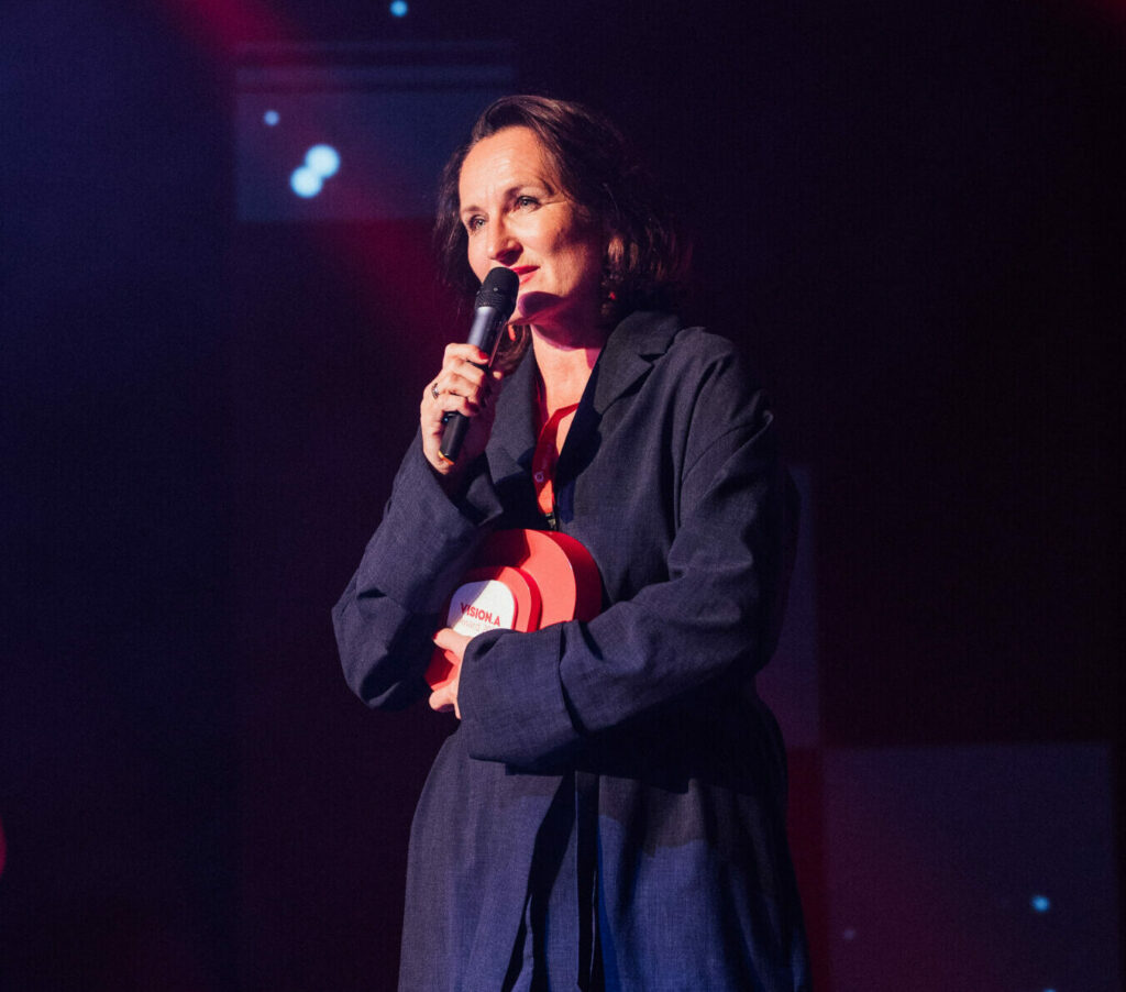 Gudrun Kreutner-Reisinger steht auf der Bühne mit dem Mikrophon in der Hand. Sie hält den roten Award und spricht.
