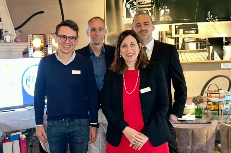 Die Gastgeber und Vortragende stehen im gemeinsamen Gruppenbild vor einem Fernseher mit Powerpoint.