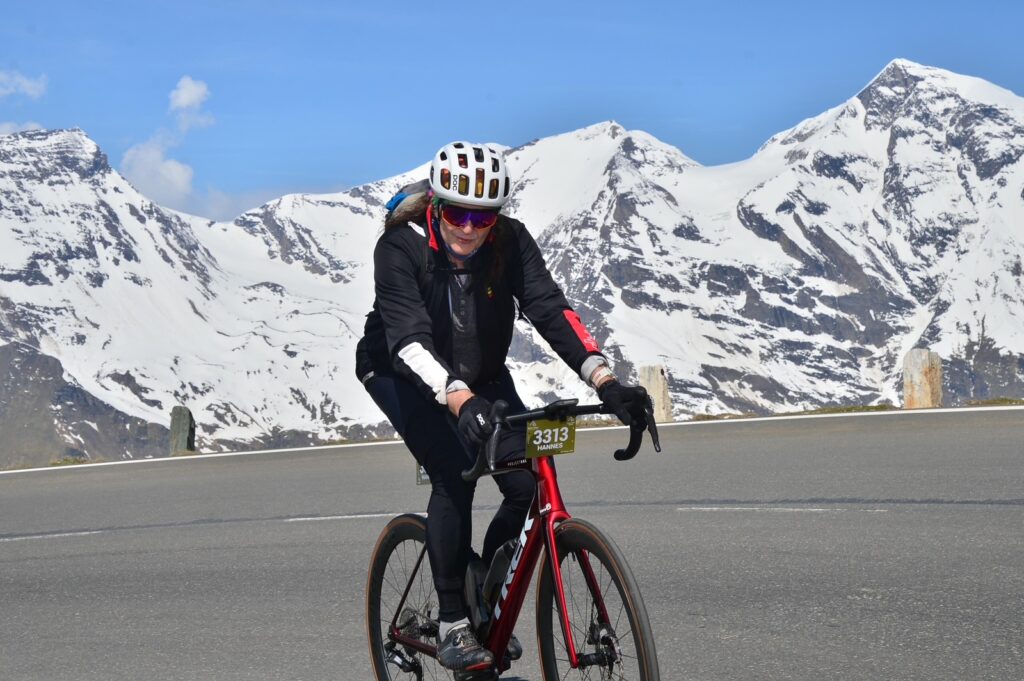 Hannes rain auf dem Mountainbike. Er fährt eine steile Straße bergauf. Hinter ihm sind die Berge zu sehen.
