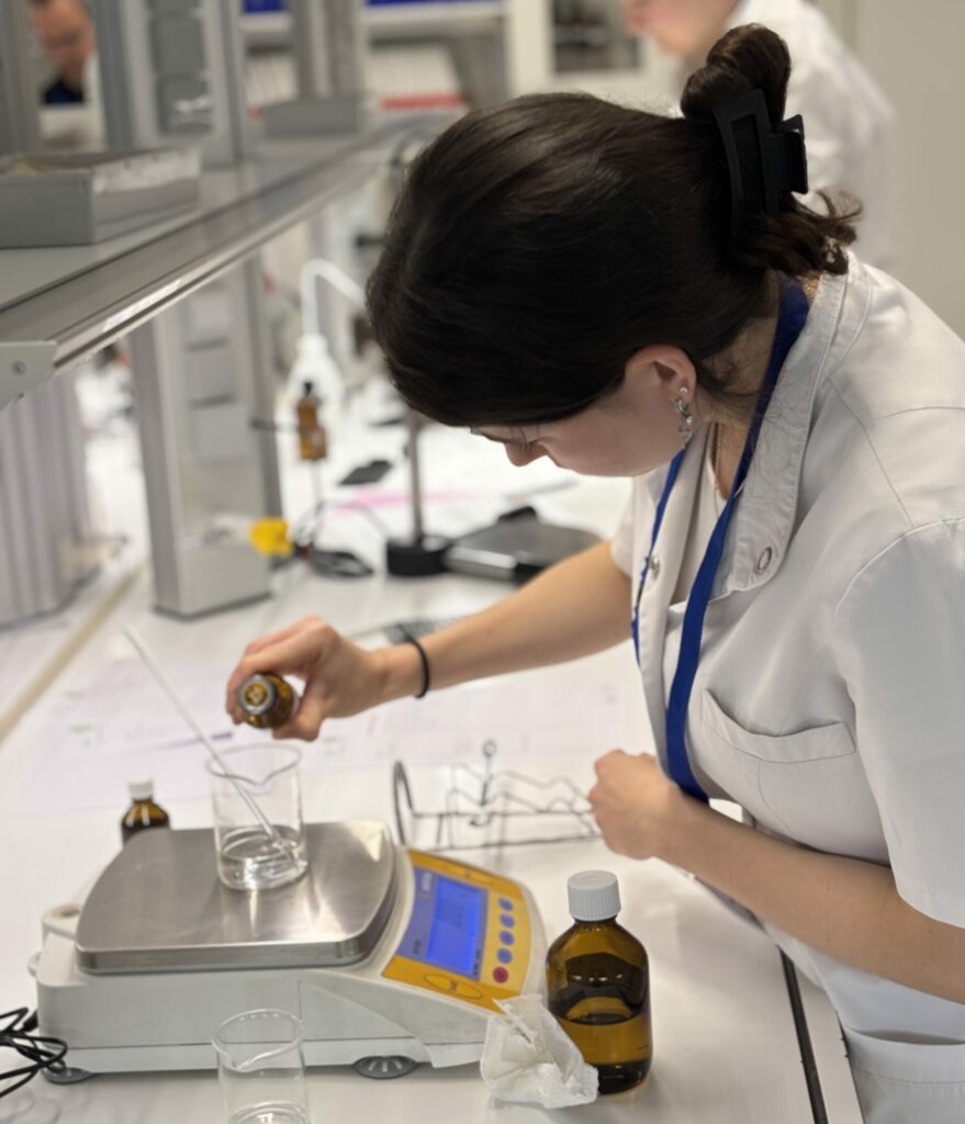 Eine Teilnehmerin wiegt eine Flüssigkeit auf einer Waage ein. Sie trägt einen weißen Mantel und arbeitet im Labor.