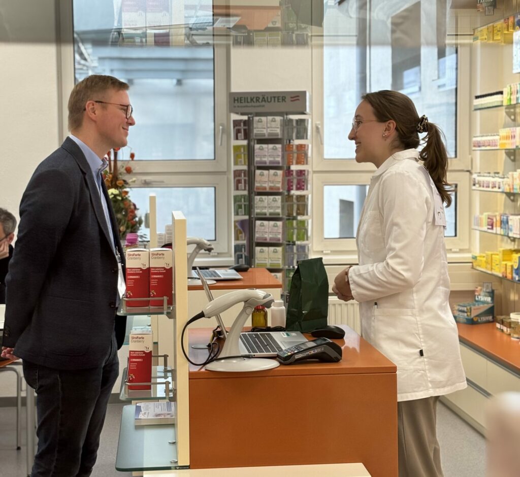 Ein Mann im Anzug wird von einem PKA-Lehrling beraten. Sie stehen an der Tara und neben ihnen sind Teeregale zu sehen.