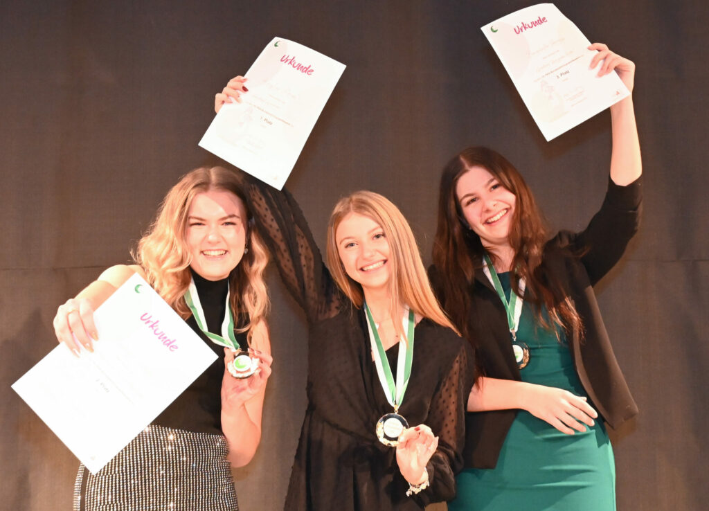 Die Top drei Gewinnerinnen halten ihre Urkunde und die Medaille in die Höhe und lachen.